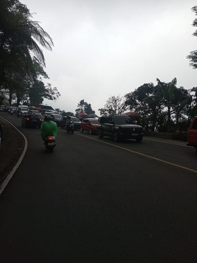 The image depicts a road in the Mae Chan District, Thailand, filled with vehicular traffic. The sky is overcast. A motorcycle, ridden by a person wearing a bright green poncho, is prominent in the foreground. In the distance, a line of cars, motorcycles, and vans can be seen. Lush greenery and trees surround the road, suggesting a mountainous or hilly location. The road is paved and marked with a yellow line.