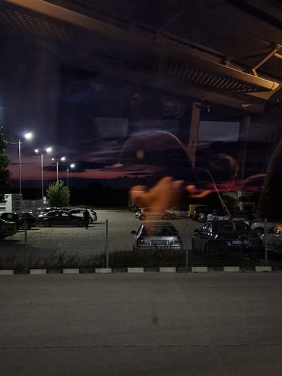 The image depicts an outdoor parking lot at dusk, with a dramatic purple and pink sky as the backdrop. Several cars are parked, including a silver Audi with license plate OB 4104 BB, and a dark-colored SUV with license plate EH 163 ME. A single individual, dressed in dark clothing, is walking in the parking lot. Streetlights illuminate the scene. The image appears to be taken from within a building, as indicated by reflections on the glass. The location is identified as a parking area in Bahovitsa, Bulgaria.
