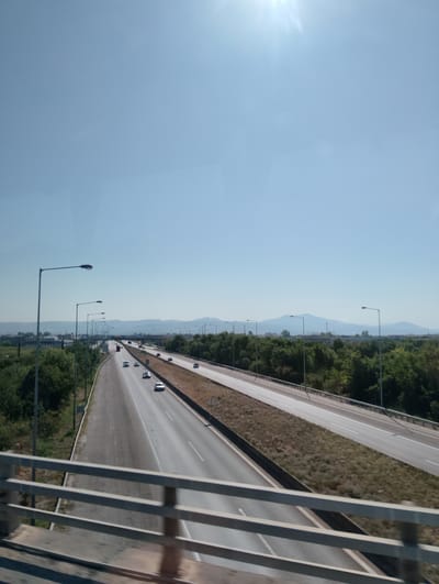 The image depicts a highway in the Municipal Unit of Echedoros, Greece, on a clear, sunny day. The perspective is from an elevated position, looking down onto a multi-lane highway with traffic. On the left side of the highway, there are lampposts and a grassy shoulder with some sparse vegetation. Beyond that, there is a line of trees and further in the distance, mountains under a clear blue sky with the sun casting a bright glare. On the right side of the highway, there is a similar arrangement of trees and distant mountains. Several cars are visible on the highway, traveling in both directions. The foreground shows a portion of a metal railing, suggesting the viewpoint is from an overpass or a bridge. The overall impression is of a modern infrastructure cutting through a natural landscape.
