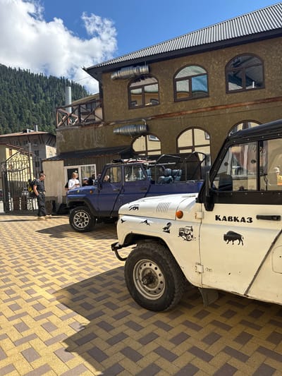 In Dombay, Russia, two off-road vehicles are parked on a cobblestone courtyard in front of a two-story building with arched windows and a metal roof. The building has a textured, tan facade, with metallic ductwork visible on the exterior. A dense, green, forested mountainside rises behind the building under a bright blue sky with scattered clouds.

The vehicle in the foreground is white with a black roof and fender flares. It has "КАВКАЗ" written on its side, along with several decals: a stylized mountain range, a wolf, an off-road vehicle, and a bison. A "4x4" decal is also present. The vehicle's front wheel and part of the side panel are visible.

Behind the white vehicle is a blue off-road vehicle, also with a black roll cage and seats. A man with a yellow backpack stands to the left of the blue vehicle, facing a group of people near the vehicle's open door. He appears to be talking to them. The courtyard is paved with interlocking yellow and grey bricks, casting shadows from the vehicles and building. To the far left, an ornate metal gate stands open, revealing a glimpse of another building and more of the cobblestone paving.