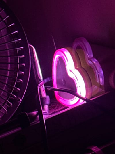 The image shows a close-up of a neon heart-shaped light that is pink and white. The light appears to be resting on a black surface, and a silver fan with a metal grate is visible to the left of the neon light. The background is dark and out of focus, with a purple hue. There are no people or discernible actions taking place. The specific location in Sant Boi de Llobregat, Spain, cannot be determined from the image itself.