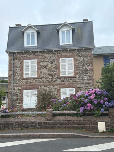 A stone-built house with a gray slate roof and dormer windows stands in La Clarté, France. The house has two stories with four white-shuttered windows on the first floor and two dormer windows on the second floor. The exterior walls are constructed of rough-hewn, reddish-brown stones. In front of the house, a low stone wall encloses a garden filled with blooming hydrangeas in shades of pink and purple. A gray metal railing runs along the top of the stone wall. A street with a pedestrian crossing is visible in the foreground. The sky is overcast.