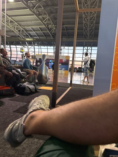 The image depicts an interior view of Zaventem Airport in Belgium. In the foreground, a person's leg and foot, wearing a light gray sneaker, are visible. The individual appears to be sitting, as their leg is bent and resting. In the mid-ground, several individuals are seated on a bench or waiting area. One man on the left is wearing a patterned shirt and dark pants. Next to him, a man in a blue and white striped long-sleeved shirt and shorts is looking to the side. Between the seated men is a large, silver cello case. Luggage, including a black and gray duffel bag and an orange and black duffel bag, are on the floor in front of them.
Further back, a man with a backpack is standing and looking towards large windows that offer a view of the sky, suggesting either dawn or dusk. The airport's interior features a high ceiling with exposed metal beams and ventilation systems. Large glass windows dominate the background, offering a view of the outdoor environment. The overall scene conveys a sense of waiting or transit within the airport terminal.