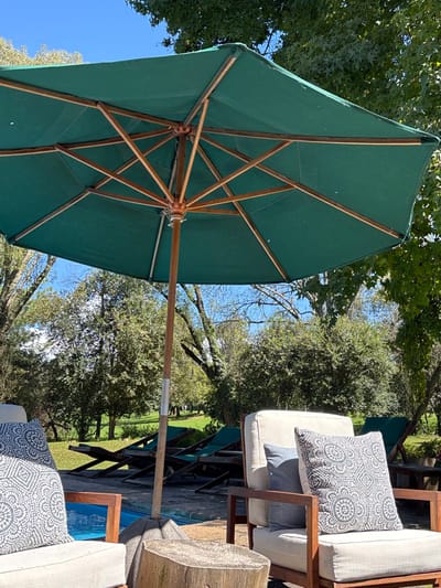 The image depicts an outdoor leisure area in Huasca de Ocampo, Mexico, under bright daylight. A large, open, hexagonal or octagonal market umbrella with a dark green fabric canopy and a visible wooden support structure (ribs and pole) occupies the upper and central portion of the frame. The umbrella's wooden pole extends down to a grey, concrete base, partially obscured by a cut tree stump serving as a rustic side table in the immediate foreground.
Two empty armchairs are visible in the foreground. The left armchair, mostly out of frame, is light-colored with visible wooden arms and features a grey cushion with a white circular patterned design. The right armchair, also light-colored with wooden arms, holds a solid grey cushion and a grey cushion with a similar white circular patterned design.
Behind the foreground elements and the umbrella pole, a swimming pool is partially visible with its blue water. Several dark green fabric lounge chairs with wooden frames are arranged around the pool's edge. The background consists of a dense, lush green landscape dominated by numerous trees and bushes under a clear blue sky, suggesting a resort, garden, or park-like setting. No individuals are present in the scene.