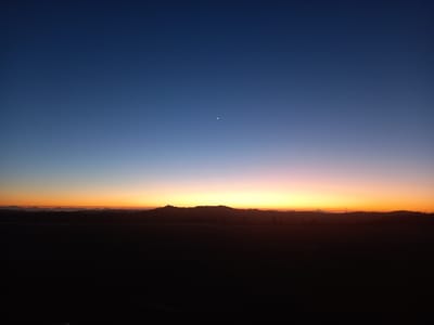 The image displays a twilight landscape over Santa Juana, Chile. The upper two-thirds of the frame consists of the sky, presenting a horizontal color gradient from deep indigo blue at the zenith, transitioning through lighter blues and purples, to vibrant oranges and yellows along the horizon. A distinct, thin white crescent moon is visible in the upper-left quadrant of the dark blue portion of the sky. Along the horizon, a dark, undulating silhouette of distant hills or forested terrain is present, contrasting with the brighter sky. The lower third of the image is occupied by a uniformly dark, undifferentiated expanse, consistent with land or water under low light conditions, offering no discernible specific details. No people, specific objects, or actions are identifiable in the foreground.