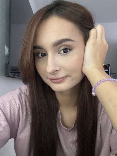 A young woman with long, dark brown hair and fair skin is captured in a close-up, selfie-style portrait. She is wearing a light pink, ribbed t-shirt and has a soft, natural makeup application. Her right hand is resting on her cheek, and she is wearing a delicate purple beaded bracelet on her left wrist. The background is blurred and appears to be an indoor setting, possibly a room with a light-colored wall. There is no indication or context provided that places this image within the city of Kuyuki, Russia.