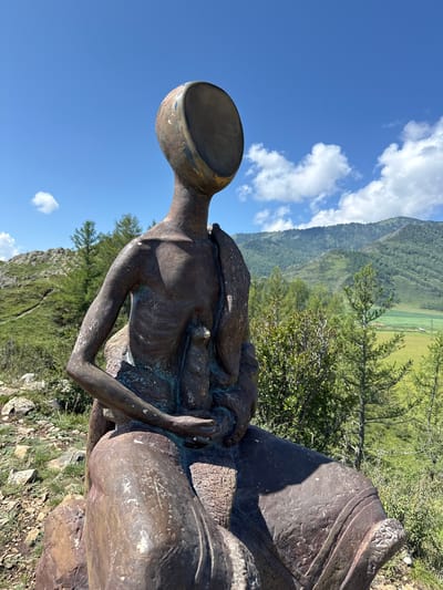 The image shows a bronze sculpture of a seated figure with an empty, bowl-like head. The sculpture is positioned outdoors, in a natural landscape. The figure is dark brown and appears to be aged, with visible texture and some green patina. It is seated on what seems to be a rough, stone base.

The background features a lush, green mountainous terrain under a bright blue sky with scattered white clouds. The foreground and middle ground show grassy slopes, rocky outcrops, and scattered evergreen trees. A winding path is visible on the left side of the image, leading up a hill. In the distance, there are more rolling hills and a field that appears to be cultivated, with some small buildings or structures.

The specific location is identified as Bichiktu-Boom, Russia. The sculpture is situated in a scenic, elevated spot offering panoramic views of the surrounding natural environment. The overall impression is one of a contemplative or symbolic artwork placed within a striking natural setting.