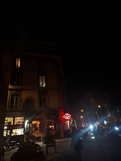 A low-angle nighttime shot shows a building with a red neon sign depicting a flower-like shape on its roof. Lights from vehicles and streetlights are visible in the background. The building has several lit windows, and an open balcony area is visible. The left side of the image is dominated by darkness.