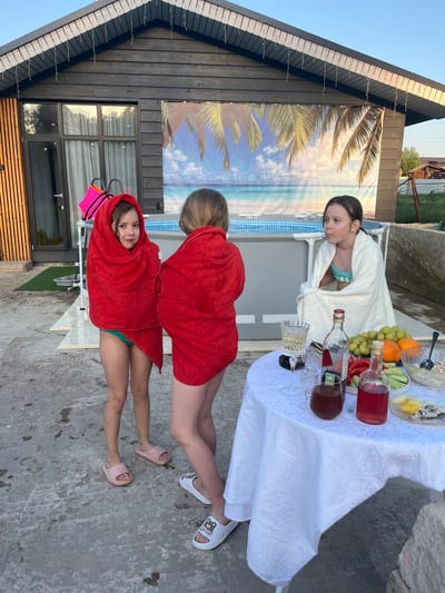 The image shows three girls standing near a pool in what appears to be the backyard of a house. Two of the girls are wrapped in red towels and wearing sandals. One girl is wearing pink sandals and the other is wearing white sandals. The third girl is wearing a white robe over a swimsuit. A table with a white tablecloth is present, with various fruits, drinks, and food items on it. Behind the girls, there is a house with a dark wood facade and a backdrop of a beach scene. The location is Новая Чернушка, Russia.