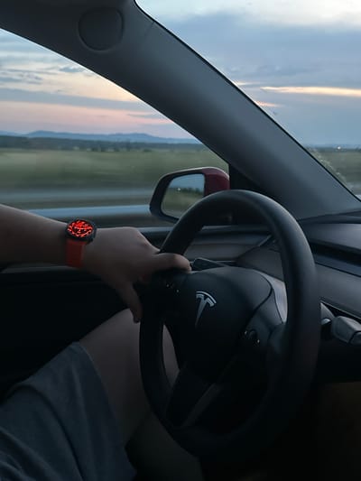 The image displays the interior of a Tesla vehicle, observed from the driver's perspective in Momkovo, Bulgaria. The driver's left hand, adorned with a watch, grips the black steering wheel featuring the prominent Tesla logo. The watch has an orange/red strap and a circular, illuminated red dial. The dial presents white numerical indicators arranged radially (e.g., 60, 100, 120, 150, 180, 200, 250, 280, 300), with a red marker pointing towards the higher end of these numbers, suggestive of a speed or RPM gauge. The driver's leg, partially visible, is covered by light-colored fabric (shorts or pants).
Through the windshield, a twilight sky is visible, characterized by soft gradients of light blue, pink, and orange hues, interspersed with scattered clouds. A distinct, hazy silhouette of distant mountains or hills spans the horizon above a flat, green and brown landscape. A portion of the vehicle's red side mirror housing is visible on the right. On the far right horizon, a small, luminous orange point of light is discernible.