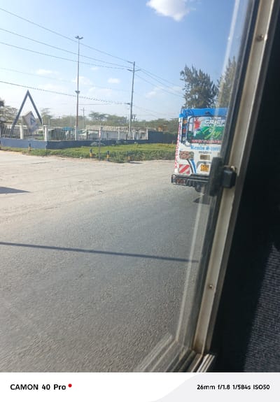 The image captures a sunny outdoor scene, likely taken from inside a moving vehicle looking out through a window. The right side of the frame is dominated by the silver-colored frame of the vehicle's window, which also casts a dark shadow diagonally across the road in the foreground.

The main subject in the midground is an asphalt road, appearing somewhat dusty or light grey. On the left side of the road, there's a dirt shoulder that transitions into a small, manicured patch of green grass adorned with yellow-and-black striped bollards. Beyond this, a fence encloses an area featuring a distinctive large, dark blue or black triangular metal structure. In the background on the left, utility poles with power lines stretch across the clear blue sky, along with some distant trees and industrial-looking buildings or containers.

On the right side of the road, the rear of a colorful public transport minibus, commonly known as a 'matatu' in Kenya, is visible. It has a blue roof and a white body with distinctive red, white, and black stripes and graphics. Text visible on the back of the matatu includes an inspirational message: "YOUR GRIEF WILL TURN INTO Joy." Below this, the name "Rembo Shuttl" and the vehicle manufacturer "ISUZU" are legible. The license plate reads "KDG 639E." A graphic of a person with raised arms is also part of the vehicle's decoration.

The sky is bright and clear with scattered white clouds, indicating good weather. The overall impression is that of a daytime journey through a semi-urban or rural area, possibly in Kenya, given the style of the matatu and license plate format.

Camera information overlaid on the bottom of the image indicates it was taken with a "CAMON 40 Pro" at "26mm f/1.8 1/584s ISO50".