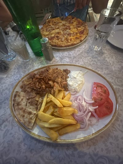 A plate of Greek food is centered on a table covered with a white patterned tablecloth, with a pizza in the background. The plate contains gyros meat, french fries, pita bread, tzatziki sauce, sliced tomatoes, and red onions. A green bottle, two glasses, and a salt shaker are also on the table. The scene is lit by overhead lighting, casting shadows on the food and table. The background is blurred, but appears to show people dining at a restaurant. The location is the Municipal Unit of Ellomenos, Greece.