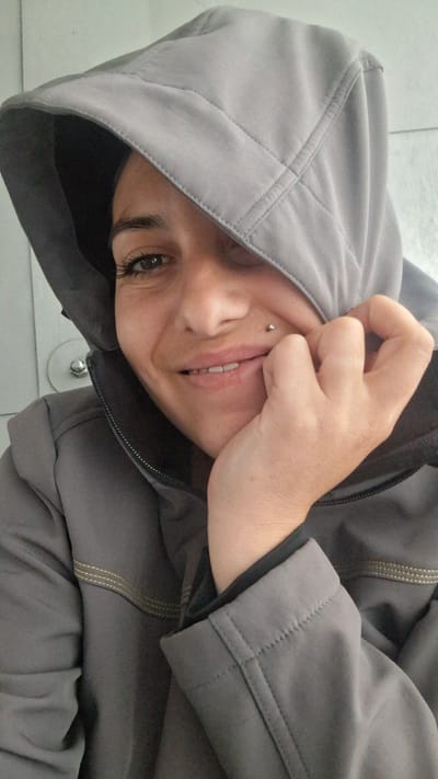 A close-up, waist-up shot shows a woman wearing a grey hooded jacket and resting her chin on her hand. Her face is turned slightly to the right, and one eye is partially obscured by the hood. She has a small silver stud piercing above her lip. The background is out of focus and appears to be a light-colored wall. The image was taken in Račice, Czechia.
