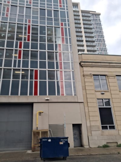 This image captures an exterior view of a modern building with distinct architectural features. The building exhibits a mix of glass and concrete elements, with a prominent facade featuring a grid of windows. Some windows are dark, reflecting the sky, while others appear to have a lighter tint. Horizontal bands of red accent the grid pattern on the lower section of the building. Above this, a taller, older building with a more classic design is visible, characterized by a series of vertical window bays and decorative stone detailing. The sky appears overcast, suggesting a cloudy day.
In the foreground, a large blue dumpster is visible on the right side of the frame. There is some text on the dumpster, though it is partially obscured and illegible. A yellow pipe extends from the building to a metallic structure beside the dumpster. The scene appears to be set in an urban environment, likely during daylight hours given the illumination. No people are visible in the image. The overall impression is one of an urban landscape with a blend of architectural styles.