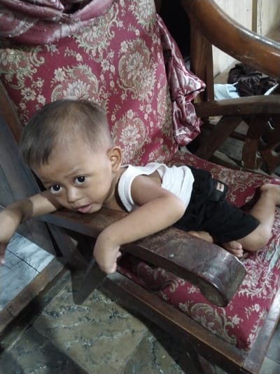 A young child, appearing to be between 1 and 2 years old, is lying on a wooden chair with a red patterned cushion. The child is wearing a white tank top and black shorts. Their head is resting on the armrest of the chair, and their arms are outstretched. The child's eyes are open and looking towards the camera. The background shows a wooden structure and some fabric. The scene appears to be indoors. The floor is tiled with a light-colored pattern. The image was taken at Winong, Indonesia.