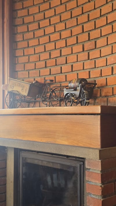 The image shows a decorative metal model of an ice cream cart and a separate cart with a barrel, positioned on a wooden mantelpiece above a fireplace. The background is a brick wall. The scene is indoors, likely in a domestic setting. The model ice cream cart is a tricycle with a large box in the front labeled "ICE CREAM" with images of ice cream cones. The other cart is a two-wheeled cart with a barrel on it. The mantelpiece is made of polished wood, and the fireplace below has a dark glass door. The overall impression is of a decorative display. The specific location in Parekklisia Community, Cyprus, is not discernible from the image itself.