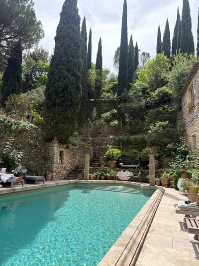 This vertically oriented image captures a serene outdoor swimming pool area within a terraced garden, likely in Saint-Victor-la-Coste, France. The scene is bathed in bright daylight, suggesting a clear or partly cloudy day with visible shadows indicating mid-day to afternoon.

The main subject is a turquoise swimming pool, reflecting sunlight off its rippling surface. The pool is bordered by light-colored stone paving. On the pool deck, a person is partially visible in the lower-left corner, relaxing on a white sun lounger and reading. Nearby, another wooden sun lounger and a dark bag are also present.

Beyond the pool, a lush, multi-level garden unfolds, characterized by extensive stone walls and steps. Numerous tall, slender cypress trees dominate the background and left side, towering against a bright sky. The garden features diverse greenery, including shrubs, climbing plants on the stone walls, and potted flowers and plants. A rustic stone building with windows is partially visible on the left side, integrated into the natural landscape.

In the middle ground, nestled within the garden, there's a small outdoor seating area with a table and chairs, some draped with light-colored fabric. A weathered stone column or structure stands prominently near this area. The overall setting evokes a sense of tranquility and a charming, natural ambiance, typical of a Mediterranean or Provençal landscape. No discernible text is visible in the image.