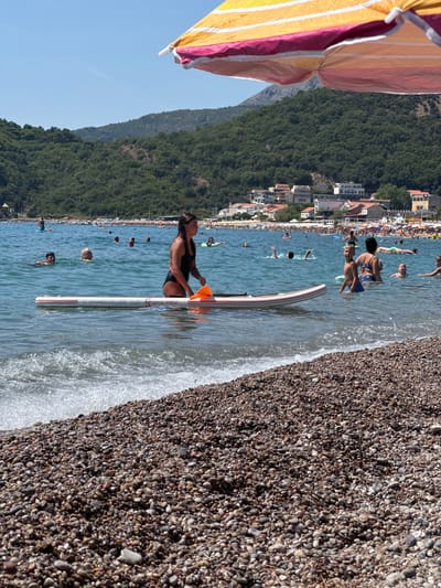 This image captures a sunny day at the beach in Đurovići, Montenegro, with the Adriatic Sea in the foreground and a lush green mountainside dotted with buildings in the background. A person in a black swimsuit is on a stand-up paddleboard, holding an orange paddle. Several other individuals are visible in the water, some swimming and others relaxing. The beach itself is composed of small, smooth pebbles. A large, colorful beach umbrella is partially visible at the top of the frame, casting a shadow over the scene. The water is a clear blue, with small waves lapping at the shore. The general atmosphere is one of leisure and summer recreation.