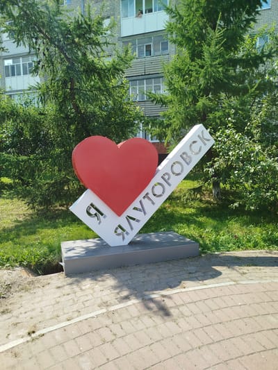 This is a medium shot, eye-level, outdoor photograph of a public art installation in Yalutorovsk, Russia. The installation features a large red heart placed on top of a white, diagonally-slanted rectangular prism. The prism has cut-out Cyrillic letters spelling "Я ЛУТОРОВСК", which translates to "I Love Yalutorovsk". The installation is situated on a grey rectangular base, which rests on a paved area. Behind the installation, there is a grassy area with trees and a multi-story brick apartment building. The building has several windows, some with balconies, and one window on the top floor shows a person standing. The trees are green and leafy, with some pine cones visible on the branches. The sky is not visible. The overall scene depicts a sunny day in a residential area.