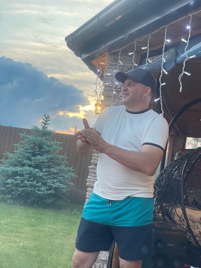 A man in a white t-shirt, black baseball cap, and teal and black shorts stands outdoors in what appears to be a backyard. He is clapping his hands. Behind him, a small evergreen tree stands in a grassy yard, bordered by a wooden fence. A string of lights hangs from the eaves of a structure behind the man, which is made of stone and wood. In the background, the sky is cloudy with hints of sunset colors. The scene is located in Chulym, Russia.