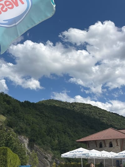 The image depicts an outdoor scene on a sunny day with a backdrop of a lush green mountain covered in trees. In the foreground, there are several white umbrellas with the "DEVIN" logo, suggesting a commercial establishment, possibly a restaurant or resort. A stone building with a tiled roof is visible behind the umbrellas. Part of a blue inflatable pool is also visible in the lower left corner. The sky is a vibrant blue with scattered white clouds. In the top left corner, a partial view of a light blue and white flag with red lettering is visible, which appears to be a promotional banner. The presence of a mountain, trees, umbrellas, and a building suggests this location is likely a resort or leisure area situated in or near Pchelishte, Bulgaria, which is known for its natural landscapes and potential for outdoor recreation. There are two men visible in the lower portion of the image, one partially visible and shirtless under an umbrella, and another seated further to the right.