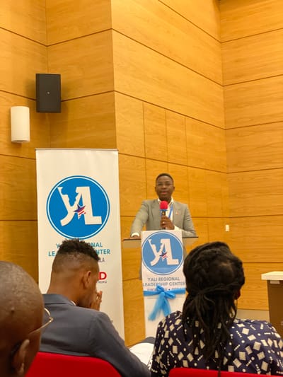 The image captures an indoor event, likely a presentation or conference, seen from an audience perspective. A man, dressed in a light-colored suit, stands behind a clear podium on the left side of the frame, speaking into a red microphone. He is addressing an unseen audience to his right.
The podium features a banner with a blue circular logo displaying a white stylized "YL" and a star. Text on this banner clearly reads "YALI REGIONAL LEADERSHIP CENTER" and "ACRP LUANDA ANGOLA". A light blue decorative ribbon is tied around the podium. Behind the podium, a larger, floor-standing banner with the same YALI logo is partially visible, also bearing the text "YALI REGIONAL LEADERSHIP CENTER" and the word "PEOPLE".
The background wall is covered with light-colored wooden panels arranged in diagonal sections. A black speaker box is mounted on this wall to the left of the speaker, and a cylindrical white object rests on the floor below it.
In the foreground, on the right side, parts of several audience members are visible from behind. One person has long dreadlocks and wears a dark patterned top. Another person is seen from the back, sporting a faded haircut and wearing a dark suit jacket. A red seat or chair back is also visible in the lower right corner. The event appears to be a formal gathering, possibly a leadership development program, taking place in Luanda, Angola, as indicated by the visible text.