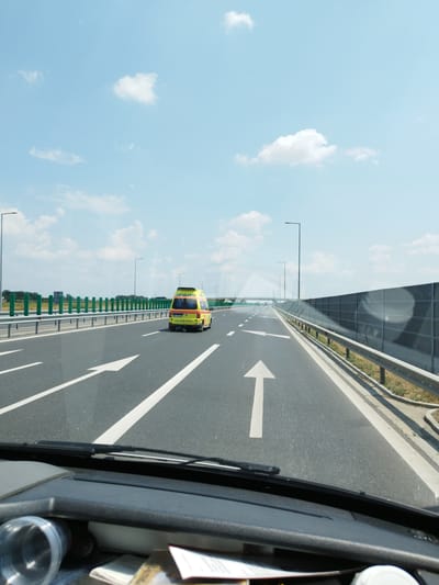 The image captures a view from inside a vehicle driving on a highway in Măgurele, Romania. The road ahead is asphalt with clear white lane markings, including a prominent upward-pointing arrow. To the right, a sound barrier wall lines the highway, and further to the left, a metal guardrail separates the road from a grassy verge with green protective netting. An ambulance, clearly marked with "Ambulanța" in yellow and red, is visible in the distance, also traveling on the highway. The sky above is a bright blue with scattered white clouds. The dashboard of the vehicle is partially visible in the foreground, showing a windshield wiper and some items on the dashboard.