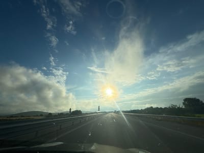 The image depicts a sun-drenched highway in the vicinity of Yazdach, Bulgaria. The perspective is from within a vehicle, looking forward down the asphalt road which stretches towards the horizon. The sky is a vibrant blue, interspersed with dramatic, wispy clouds, some of which are illuminated by the brilliant sun. The sun itself is a focal point, its rays creating a strong lens flare that beams across the scene. Guardrails line both sides of the highway, and a few road signs are visible. In the distance, rolling hills covered in greenery and trees form the landscape. The overall impression is that of a journey on a clear, bright day.
