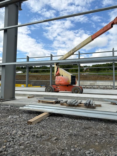 In Wilkowisko, Poland, a construction site is depicted with an orange and yellow boom lift positioned on a concrete slab. The boom lift is partially obscured by a large metal beam forming part of a building's structure. In the foreground, several piles of metal components are laid out on wooden pallets and directly on the gravel ground. The scene is outdoors under a bright, cloudy sky, with a backdrop of trees and a gently sloping hill. The construction area appears to be in an early stage, with exposed beams and foundational elements.
