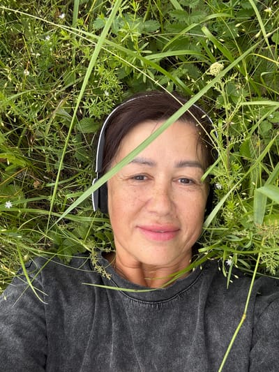 A woman with dark hair is lying on her back in the grass, wearing black headphones and a dark grey t-shirt. She is looking up at the camera, and her face is partially obscured by tall green grass. Her skin has freckles. The environment is natural, with various types of grass and small white flowers. The scene appears to be outdoors in a grassy area, possibly in the city of Kalinino, Russia, given the provided context.