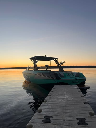 A turquoise and black wake boat with the "ATX" logo is docked at a modular floating dock on a calm body of water. The boat is positioned at an angle, with its stern facing towards the viewer. A wakeboard tower is erected on the boat, equipped with board racks. The scene is illuminated by the soft, warm glow of a sunset. The sky transitions from a pale blue at the top to a vibrant orange near the horizon, where a silhouetted tree line marks the distant shore. The water reflects the colors of the sunset and the boat. The dock is constructed from interlocking plastic modules and is wet, with some water droplets visible. The location is identified as Gromovo, Russia, suggesting a lakeside or river setting within the city. There are no people or animals visible in the image.