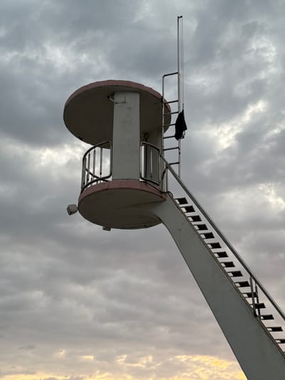 A concrete lifeguard tower with a pink trim stands against a cloudy sky in the Vardane Microdistrict, Russia. The tower has a circular platform at the top with a railing and a small enclosed area. A metal ladder extends from the platform upwards, with a black piece of fabric draped over it. A long, slanted staircase with metal railings leads up to the platform. The bottom of the image shows a sliver of yellow light on the horizon, suggesting either sunrise or sunset. The scene is devoid of people and activity.