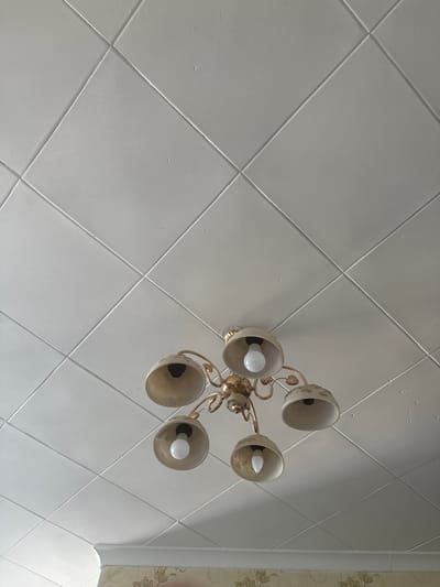 The image shows a close-up, low-angle shot of a white, tiled ceiling with a gold chandelier hanging in the center. The ceiling is divided into diamond-shaped tiles by recessed lines. The chandelier has five arms, each with a beige-colored lampshade and a visible light bulb. The top of the image is dominated by the ceiling, with the chandelier occupying the lower half. The lighting suggests an indoor environment, likely a room in a residential building. There are no people visible in the image. The location in Aqköl, Kazakhstan is not discernible from the visual information in the photograph.