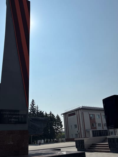 The image depicts a view of a monument and a building in the city of Belovo, Russia. In the foreground to the left is a tall, dark gray monument with red stripes. Below the stripes, an inscription in Russian reads "ГЕРОЯМ-БЕЛОВЧАНАМ ОТ ТРУДЯЩИХСЯ ГОРОДА," which translates to "To the heroes of Belovo from the city's workers." Behind the monument, a row of evergreen trees stands against a clear, bright blue sky. To the right, a modern building with a light-colored facade and red vertical accents is visible. The building has multiple windows and air conditioning units. In front of the building, there are steps leading up to what appears to be another part of the memorial. The ground is paved. A single person is visible in the distance walking on the paved area between the monument and the trees. The sun is shining brightly, creating some glare in the upper portion of the image.