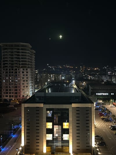 The image presents a nighttime urban scene in Skopje, North Macedonia, viewed from an elevated perspective. Dominating the upper center of the dark sky is the distinctly illuminated Millennium Cross, located atop Vodno Mountain.
In the midground, a prominent, multi-story modern building, identified as Capital Plaza, is central to the composition. It features a dark-toned facade with vertical light-colored panels on its sides and a large, central glass section with multiple illuminated interior windows. To the left of Capital Plaza stands a taller, unfinished or under-construction high-rise building, primarily concrete, with numerous exposed floor slabs and window openings.
At ground level, a paved area with several parked passenger vehicles (including red, yellow, grey, and black cars) is visible to the right of Capital Plaza, bordering a street with white markings, including a zebra crossing. To the left of Capital Plaza, a pathway or road segment is illuminated by streetlights emitting a blue light. The background features a sprawling cityscape with numerous distant lights from buildings and other urban infrastructure, extending onto hillsides. No people or specific interactions are discernible. The scene captures an urban environment under artificial illumination during nighttime.
