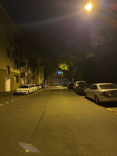 An eye-level, full shot captures a dimly lit street in Duisburg, Germany at night. A row of parked cars lines the right side of the asphalt road, illuminated by a streetlamp and ambient light. The buildings on the left are dark, with some windows faintly lit, suggesting occupancy. Trees with dark foliage are visible, casting shadows on the street and buildings. The overall atmosphere is quiet and deserted.