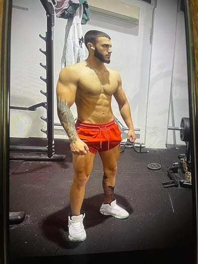 A muscular man is photographed in a home gym. He is wearing red shorts and white sneakers, and has tattoos on his arm and leg. The gym contains equipment such as a barbell, weight plates, and a rack. The room has white walls on one side and dark rubber flooring on the other. The location is not discernible as Krumovgrad, Bulgaria from the image alone.