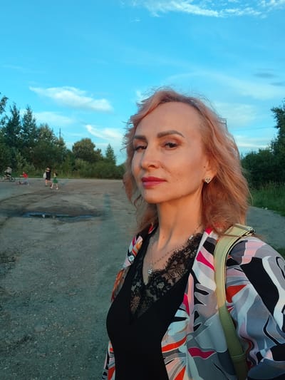 A woman with pinkish-blonde hair and makeup stands in the foreground, looking to her right. She is wearing a black top with lace trim, a colorful, patterned jacket, and a light green backpack. She has a heart-shaped earring in her left ear and a silver necklace. In the background, there is a dirt or gravel surface with some puddles. Several people are visible in the distance. One person is on a scooter, another is walking a child, and another is pushing a bicycle. Trees and foliage line the background, and a blue sky with scattered clouds is overhead. The image appears to have been taken outdoors during daylight hours, possibly in the late afternoon or early evening due to the warm lighting. The location is identified as Vasil'ki, Russia.