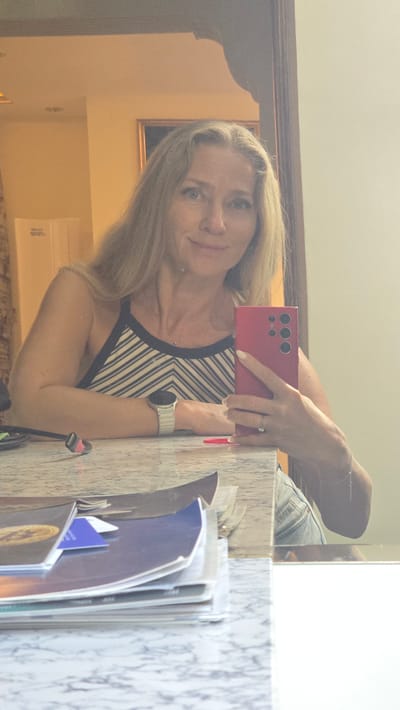 A woman with long blonde hair is taking a selfie in a mirror. She is wearing a striped tank top and a silver watch. She is holding a pink smartphone in her right hand, and a ring is visible on her left hand. In front of her, on a marble countertop, is a stack of magazines and papers. The background shows a wall with a framed picture and a doorway. The scene appears to be indoors, possibly in a hotel room or a home. The location is identified as Trikomo, Northern Cyprus / Cyprus.