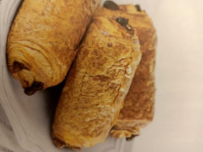 Three chocolate croissants are arranged on a white, textured surface, possibly a tablecloth or linen. The croissants are golden brown with visible flaky layers. The croissant on the left is partially obscured by the one in the center. The central croissant is in sharp focus, displaying a cracked, textured surface. The croissant on the right is slightly out of focus and positioned behind the central one. The background is a plain, light color, suggesting an indoor setting. No people or specific landmarks are visible.