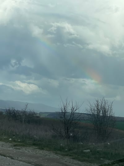 The image depicts a landscape with a prominent rainbow in the cloudy sky. To the right, there is sparse vegetation, including bare bushes. A field is visible in the distance. The ground appears to be covered in gravel. The location is Belomortsi, Bulgaria.