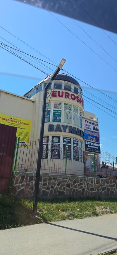 The image shows the exterior of a building in Karakol City, Kyrgyzstan. The building has a curved facade with signs for "EUROSH," "BAYMAN," and "BAYMANAP barbershop." There are also advertisements for "Yandex Taxi" and "TAKSOPARK TALK" in Russian. The building is surrounded by a fence with stone pillars. The sky is clear and blue, and there are power lines running across the top of the image. The ground is covered in grass and pavement.