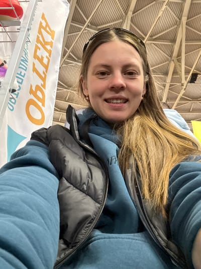 A young woman smiles warmly for a selfie, positioned close to the camera. She has long, light brown hair, a bright expression, and is wearing a blue hooded sweatshirt underneath a dark, possibly black or dark gray, puffer vest. A dark headband or pair of sunglasses rests on top of her head.
The setting is indoors, likely a large public space such as an exhibition hall or arena in Песчаные Ковали, Russia. The background is dominated by a high, light-colored ceiling with a complex geometric pattern of beams and corrugated panels. Below the woman, a white banner is visible, displaying large orange Cyrillic text that, when oriented correctly, reads "ОБЪЕКТ" (meaning "Object" or "Facility"). Below this, smaller text, partially obscured, appears to include "УНИВЕРМАГ" (Department Store). A light blue wave pattern is also visible on the banner. Some items, including a red and white object and a purple and white fluffy item, can be seen on a display rack or shelf just below the banner. The overall impression is that of a person enjoying an event or outing in a spacious venue.