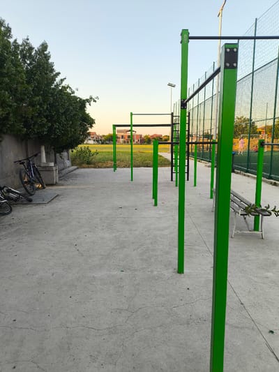 This media file depicts an outdoor park with exercise equipment, specifically a calisthenics park. The scene is set in Войводино, Bulgaria, under a clear, pale blue sky, suggesting it is either early morning or late afternoon due to the soft, warm light bathing the scene.
The primary subjects are the various pieces of green-painted metal exercise equipment, including pull-up bars and parallel bars, arranged on a concrete ground. A weathered wooden bench is also visible, with a branch of leaves resting on it. To the left of the frame, several bicycles are propped against a concrete wall, suggesting recent use or storage. In the background, behind a wire mesh fence, a grassy field and residential buildings can be seen. There are a few people in the background on the field, one wearing pink, but they are indistinct.
The setting is an open-air public space designed for fitness and recreation. The concrete ground is cracked in places, indicating some wear and tear. The overall mood is peaceful and tranquil, amplified by the calm weather and the gentle lighting. There is no discernible activity taking place in the immediate foreground; the scene appears static and ready for use. A subtle detail of text is visible on one of the upright posts of the calisthenics equipment, reading "HAYOS".