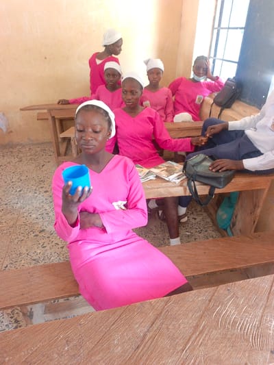 This image captures a group of individuals, predominantly young females, seated in what appears to be a classroom in Mada, Nigeria. Seven females are visible, six wearing matching long-sleeved, V-neck pink dresses and white head coverings. One male is partially visible on the right, wearing a white collared shirt and dark trousers.

In the foreground, a young female, also in a pink dress and white head covering, sits on a wooden bench, holding a small, light blue plastic cup in her right hand. She is looking down towards the cup. Her left arm is resting across her abdomen.

Behind her, five other females are seated at simple wooden desks and benches. One female directly behind her also wears the pink dress and white head covering, looking towards the left. To her right, another female in similar attire looks directly forward. A third female in pink and white head covering is seated further back, looking directly forward. To her right, a fourth female in pink and white head covering, wearing a light blue face mask pulled below her chin, leans back against the wall, resting her right arm on the desk. A fifth female stands in the background on the left, also in a pink dress and white head covering, looking right.

On the desk in the middle ground, several books or papers are stacked. A black sling bag rests on the desk next to the female in the face mask. Below the desk, a turquoise bag is visible on the floor. The male on the far right has his hands resting on the desk, one hand appearing to interact with a female's hand.

The classroom features light-colored walls, possibly off-white or light yellow, with some faint markings. The floor is covered with patterned, light-colored tiles. A window with dark frames is visible on the right, admitting natural light into the room. The wooden desks and benches appear aged and utilitarian.
