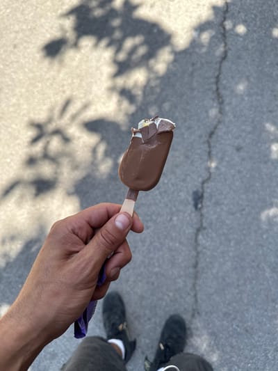 The image shows a hand holding a partially eaten chocolate-covered ice cream bar. The hand is male, with visible veins and some light body hair on the forearm. The ice cream bar is dripping, indicating it is melting in the warmth. A crumpled purple wrapper is also visible, held loosely in the hand.
The setting appears to be outdoors, with a cracked asphalt surface visible in the background. Shadows of leaves and branches are cast on the ground, suggesting the presence of trees or other foliage nearby. The lighting implies it is daytime, likely a sunny day due to the distinct shadows and bright illumination. The overall atmosphere is casual and relaxed, possibly suggesting a moment of enjoyment during a warm day.
In the background, a person's feet are visible, wearing dark shoes and grey pants, with white socks. This, along with the shadows, hints at another person being present. The location context of l'Ametlla del Vallès, Spain, further suggests a warm climate. No visible text is present on the ice cream bar itself, though the wrapper might contain some, it is not clearly legible in the image.