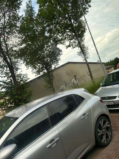 This is a photograph of a mural on a building in the city of Limanowa, Poland. The mural depicts two men in historical attire, and text in Polish can be seen above them, part of which reads "ZWIĄZEK ARMII KRAJOWEJ W LIMANOWEJ". Two silver cars are parked in front of the building, one in the foreground and another partially visible to the right. Green trees with dense foliage are present on the left side of the image, and the sky is cloudy. The image appears to have been taken from a slightly elevated position, possibly from another vehicle.