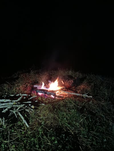 A campfire burns brightly at night in a grassy area. The flames are a vibrant orange and yellow, illuminating the surrounding dry grass and a pile of logs. The scene is dark with the exception of the fire and its glow. The location is outdoors, in what appears to be a rural or natural setting. There are no people or animals visible in the image. The fire is contained within a small area of the grass.