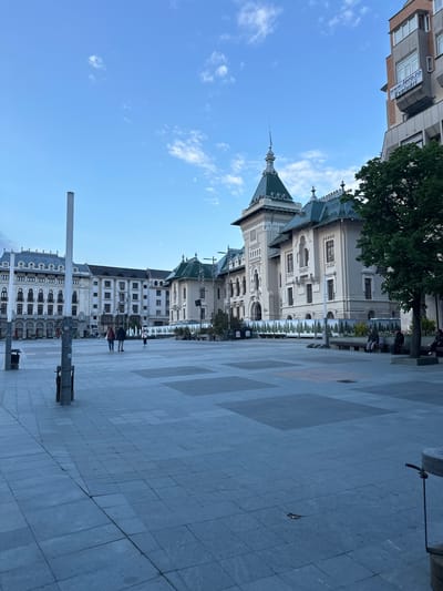 The image shows a view of Union Square in Craiova, Romania. The Prefecture building, with its ornate architecture and green roof, dominates the left side of the frame. A section of another building is visible in the upper-left corner. The foreground is a paved square with a pattern of gray and beige stones. A lamp post is visible on the bottom-left. Several people are present, mainly on the left side of the image. The sky is blue with sparse clouds. A tree is located next to the Prefecture building.