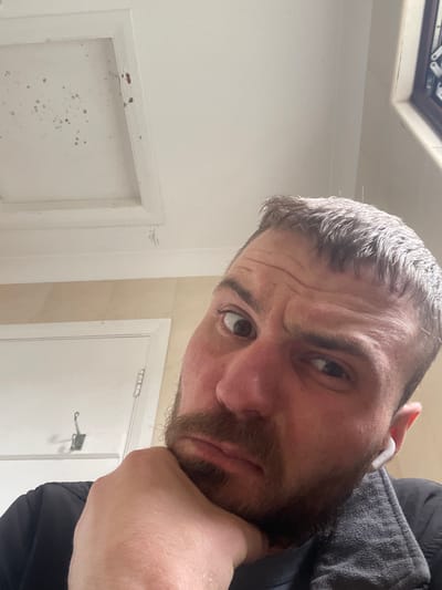 The image shows a close-up, low-angle shot of a man's face. He has short, dark hair and a beard, and is wearing white wireless earbuds. His left hand is propped up to his chin, and he appears to be looking directly at the camera with a slightly furrowed brow. The background is out of focus but appears to be an indoor setting with a white ceiling that has some marks on it, and a wall with a light brown textured finish. There is also a white door visible on the left side of the frame. The lighting suggests an indoor environment, possibly a bathroom or a hallway. No specific city or location in Ireland can be identified from the visual information presented.